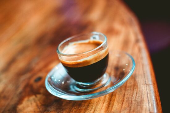 A cafecito in a clear glass espresso cup and saucer sits atop a wooden surface.