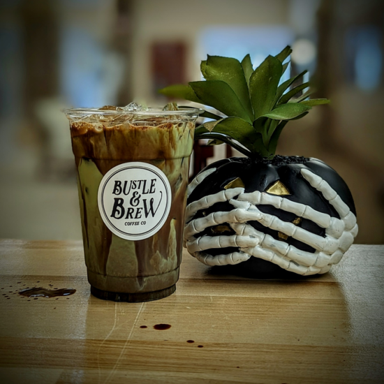 A dark chocolate iced matcha drink. Next to the matcha is Halloween decor, skeleton hands wrapped around a pumpkin.