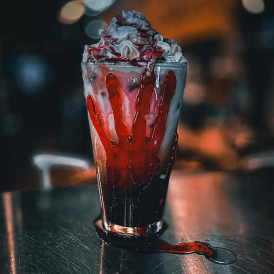 An iced drink with chocolate and cherry syrup topped with whipped cream. A creepy red gummy hand is plastered to the side of the glass, and cherry blood oozes onto the table. 