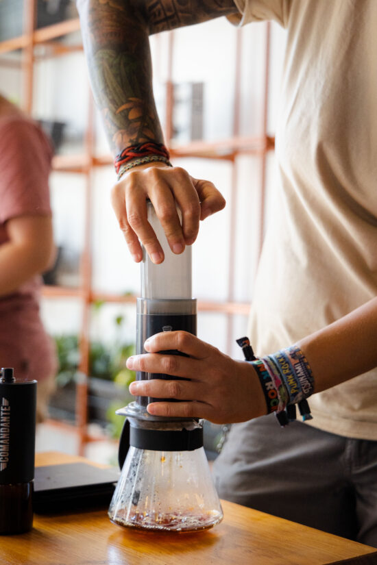 Someone pushes down the plunger of an AeroPress to brew coffee for the competition. They are wearing a white t shirt and stacked bracelets.