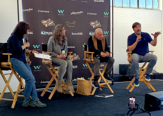 Four panelists, two women and two men, in the Leading Coffee Pioneers panel. Each sits in a director's chair on the stage in front of a blue backdrop with festival sponsors' company names printed on it. Each holds a microphone.