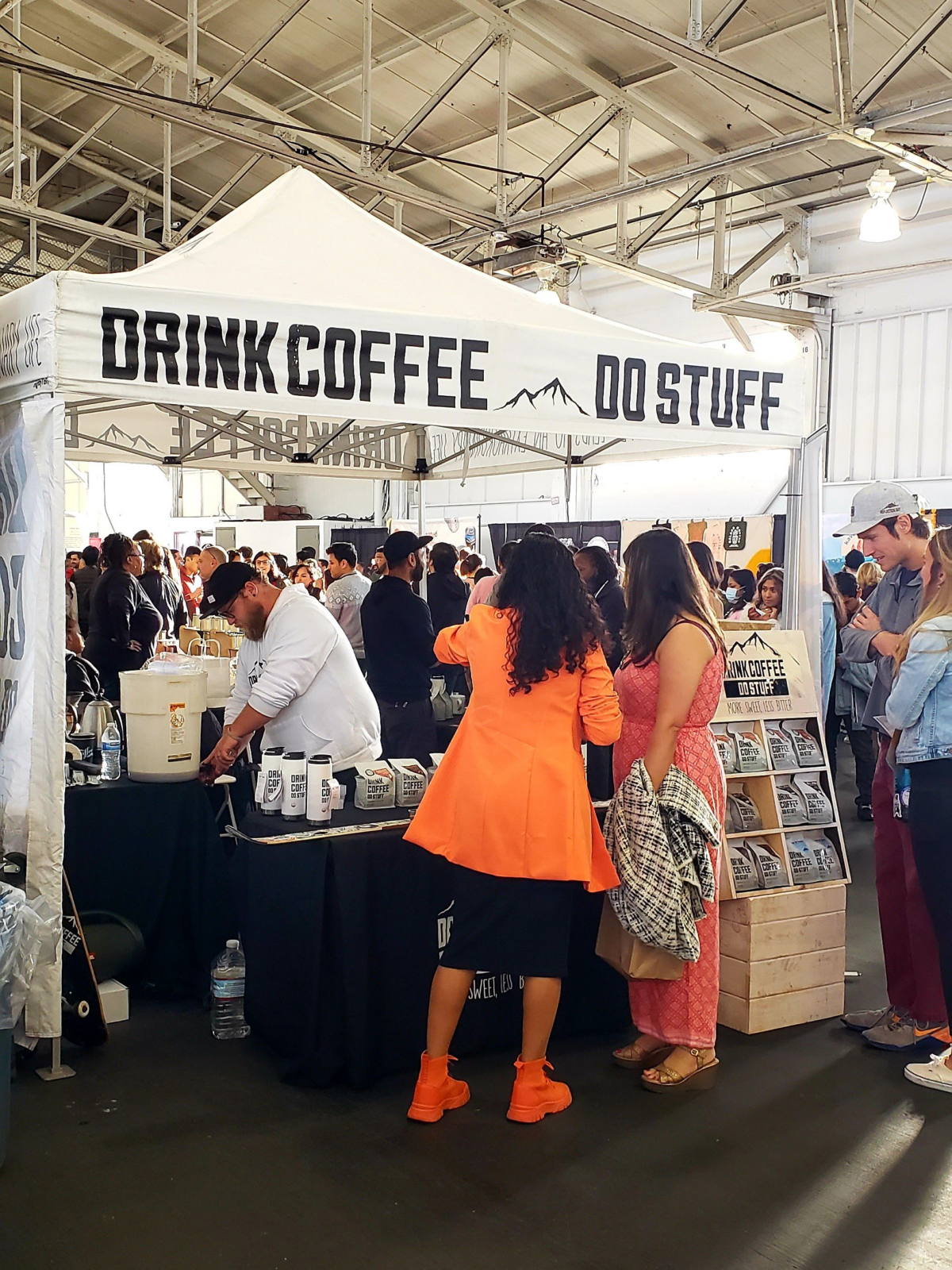 Festival goers surround a shite tent in the pavilion for the company Drink Coffee Do Stuff. They have merch like coffee bags and portable mugs on display. A bearded barista in a black hat pours cold brew for a guest from a Toddy bucket.