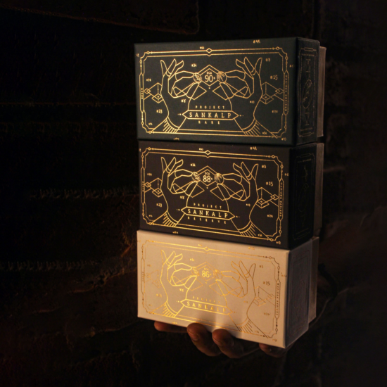 A stack of three Sankalp boxes, about the size of tea boxes, that depict illustrations of two women's hands in gold leaf. Each hand holds a large coffee bean. The hands have bracelets, and there is a border and background of minimal geometric shapes like diamonds.