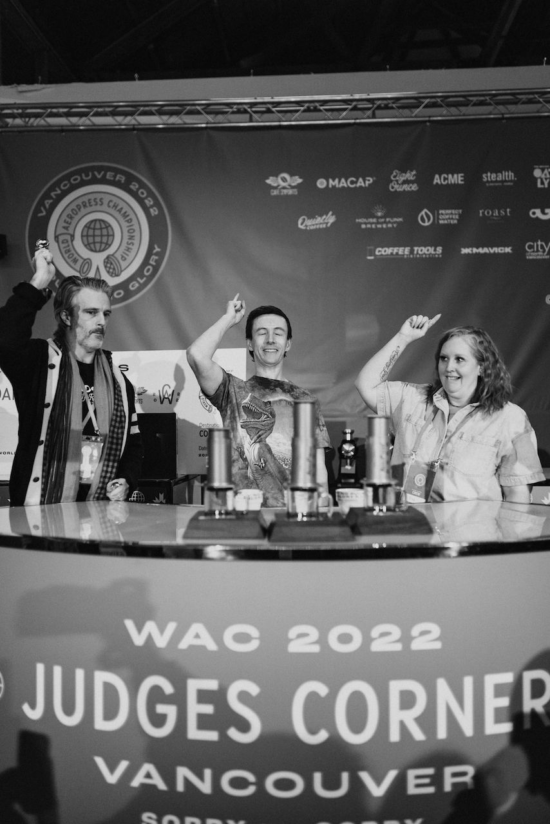 Three judges have hands with pointer fingers raised above three coffee selections, about to point to their chosen recipe. 