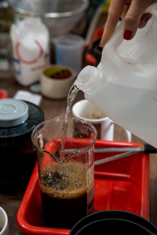 A hand with red nail polish pours clear liquid into a glass beaker with coffee grounds inside. The beaker is inside a shallow red plastic tub. Arranged on the table is other photography developing equipment and a coffee mug.