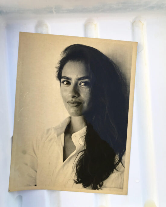 A sepia-toned portrait photo of a young woman with dark brows and long hair, smirking directly at the camera. She wears a white button-up shirt.