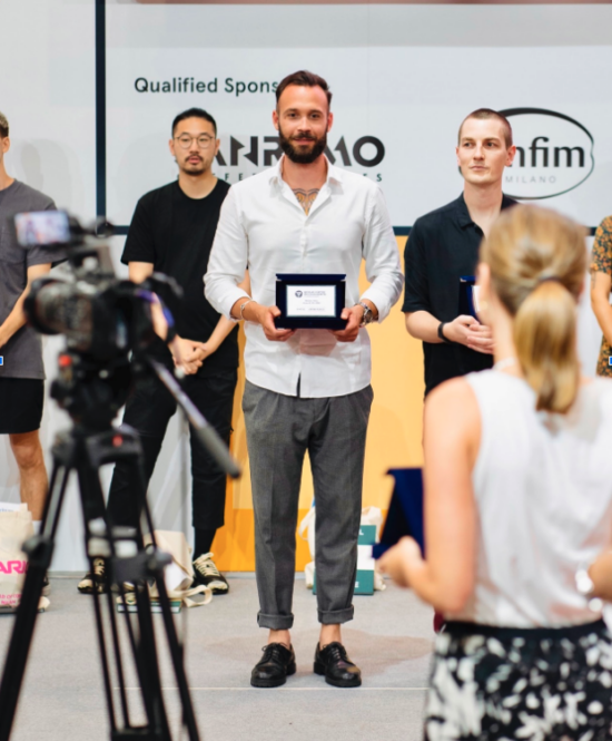 Christos stands in front of a camera with other winners at the Hellenic Coffee in Good Spirits Championship. He has a beard and short hair, and wears a white shirt, gray pants, and leather shoes. He is holding up his award, which is a black and white framed certificate. 