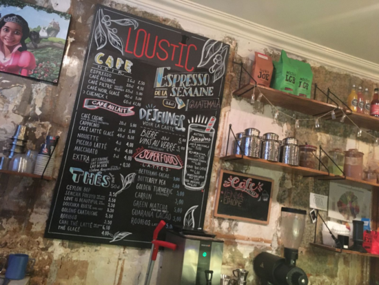 The wall behind the espresso bar at Loustic. There is a blackboard with the menu written in bright colors, shelves built into the wall to hold coffee bags and jars, a flavor wheel and other paraphernalia.