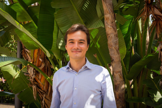 Portrait of Andrea standing before giant tropical leaves. He wears a button-up shirt and smiles.