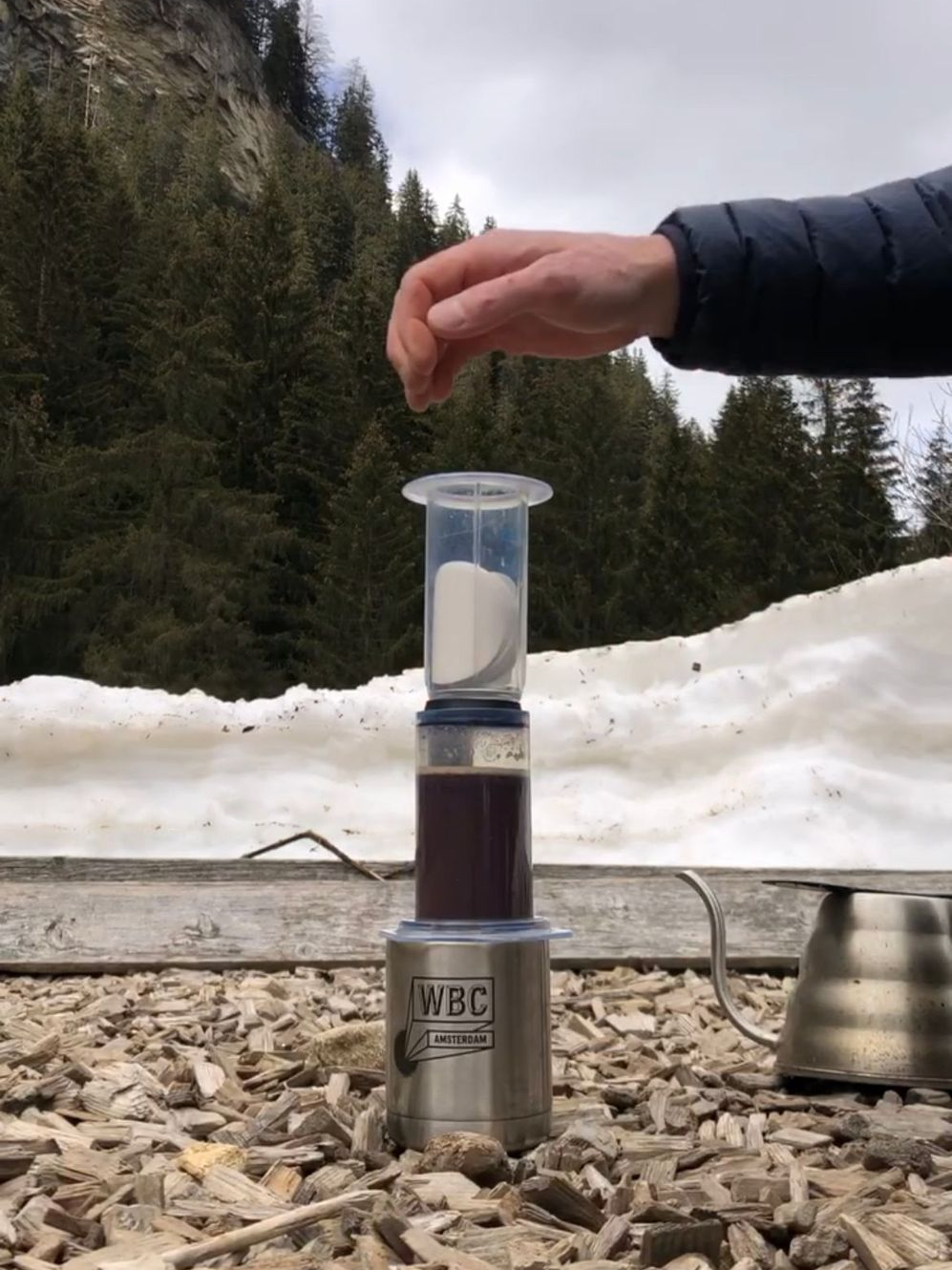 Lex has an AeroPress with coffee in it, laid on top of a metal travel mug, ready to brew. There is snow on the ground, and evergreen trees behind.