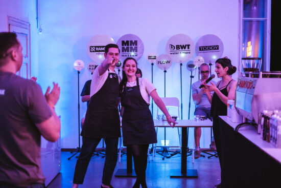 The Barista League in Budapest. Two baristas smile at the camera; one is pointing at it directly. The MC speaks into the microphone off to the side.