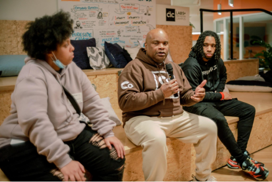 Keith sits with two teens on a wooden platform. He holds a microphone, giving a talk. He wears a brown COCC hoodie and white pants and sneakers.