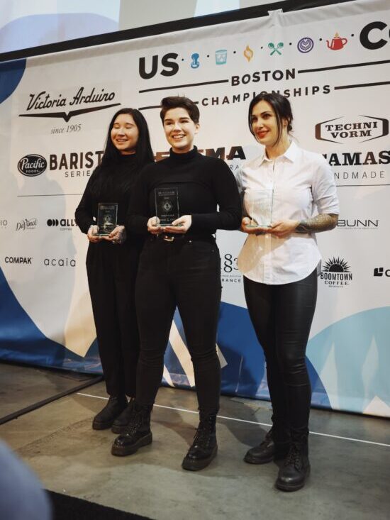 Morgan stands between two other competitors, all wearing black and white, and boots. Behind them is the Boston Championships banner with USCC logo and logos of the event sponsors.