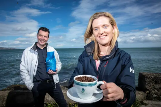 Marguerite holds up a cup and saucer while David looks on with a coffee bag in front of Clew Bay.