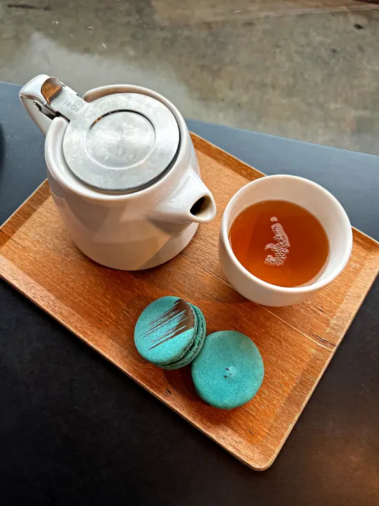 A wooden tray at Alchemist holds a white teapot with a small spout and metal lid. There is a wmite ceramic tea cup on the tray next to two small blue-green macaron cookies.