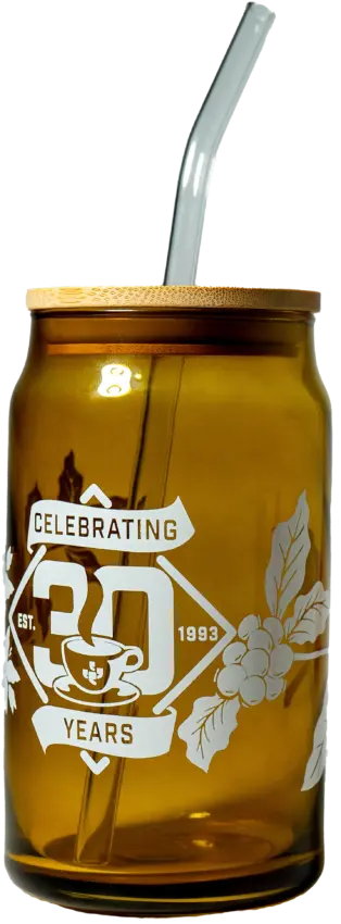 An amber colored can-shaped glass with Celebrating Thirty Years printed on the side. There are coffee plants and a coffee mug printed on the side as well. The glass has a plain bamboo lid, and clear glass straw inserted.