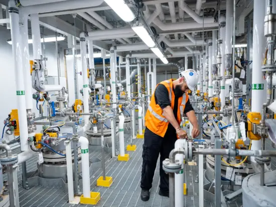 An engineer adjusts something on an intricate setup of hundreds of tubes and columns in the processing plant. He wears a hardhat and orange vest.