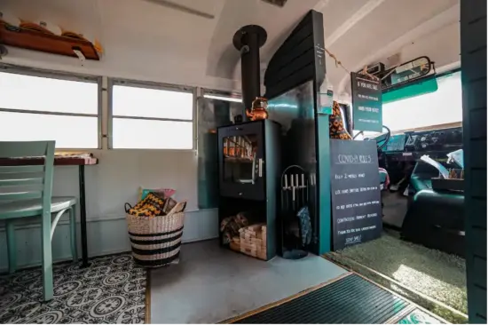 The front corner of the bus features a small metal wood-burning fireplace with a skinny chimney. Wood is piled neatly underneath. A black striped basket lies on the floor near it. Behind the fireplace is the orginal bus driver's seat and signage.