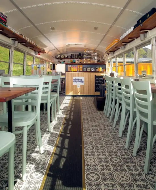 The interior of the bus: the floor is covered in mosaic style tiling, with the classic black rubber strip running through the middle. The ceiling is curved and painted white. At the end is a short wooden bar. On the left side is a row of two-person wooden tables with white chairs. On the right side runs a long skinny wooden bar seating area with the same shite chairs. The original bus windows line the top on both sides.