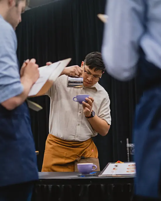 Flook in an off white shirt, glasses, and tan apron pours latte art in a purple mug. Judges in blue aprons with clipboards watch and score his performance.
