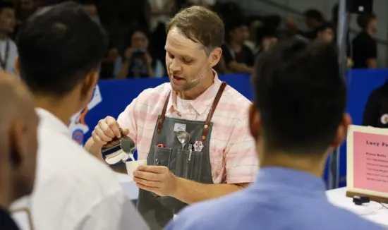 Isaiah wears a pink striped shirt with a denim apron and pours steamed milk into a ceramic cup during the competition.