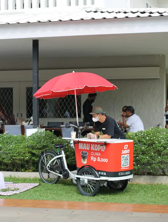 A man stands by an electric bike cart with a red umbrella. It has a picture of a coffee cup on it.