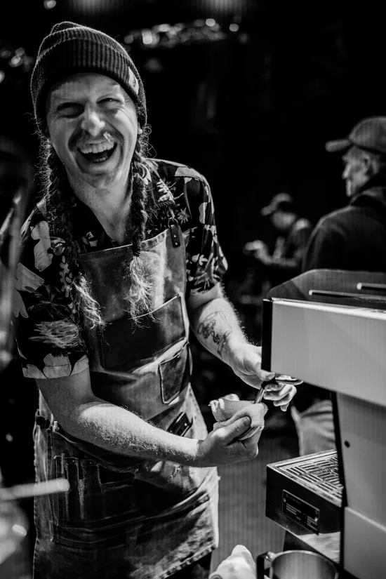 A man with braids and an apron cleans an espresso machine wand. 