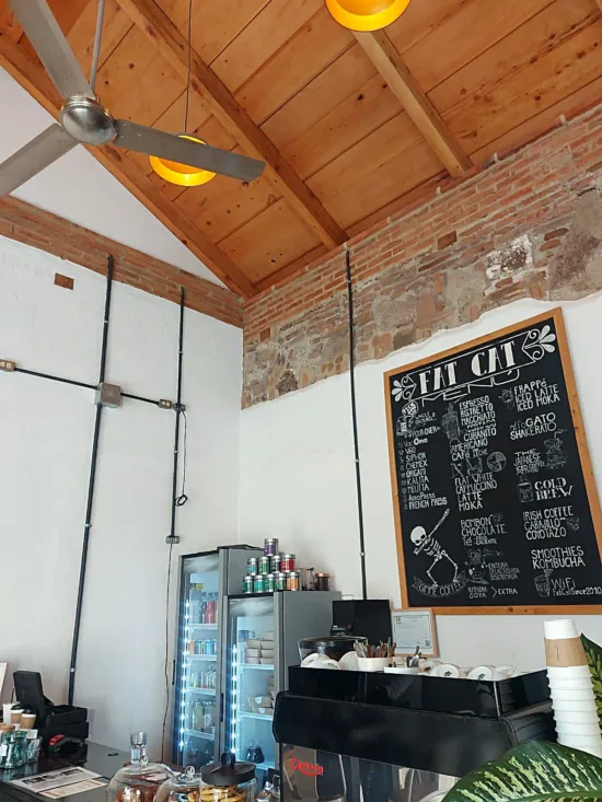 behind the bar at Fat cat, there's a chalkboard menu with a drawing of a skeleton "dapping" and drinks listed. Two small fridges are in the corner. On the bar is an espresso machine with cups stacked on top.