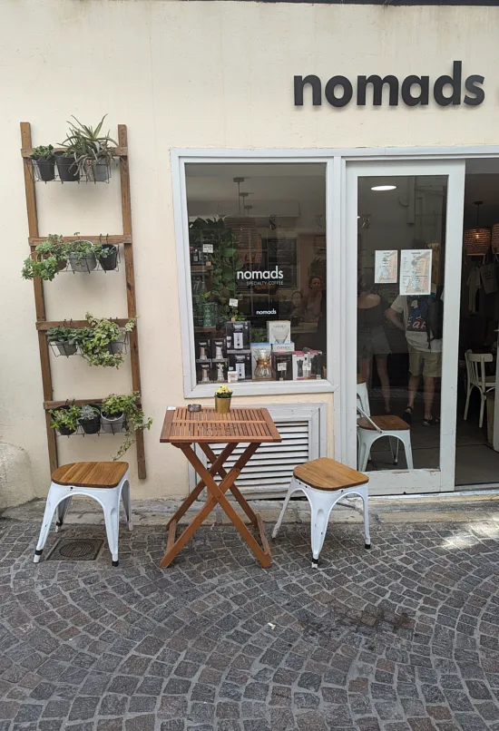 The cobblestone street outside nomads. A small wooden table with two stools sits outside. A ladder-style shelf unit on the outside wall holds small plants. Large windows look inside the cafe.