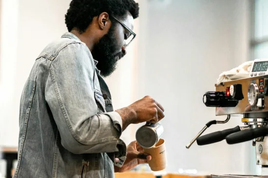 A barista pours milk from a steaming pitcher into a paper cup.