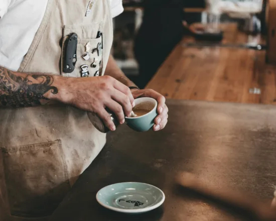 Person in apron pouring latte art in a small blue cup with matching saucer.